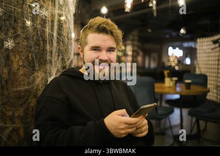 Gutaussehender, konzentrierter Kerl, der Geldtransaktionen über die Anwendung auf dem Handy macht, mit 4g Internet-Café am Straßenabend, ernsthaft Millennial Stockfoto