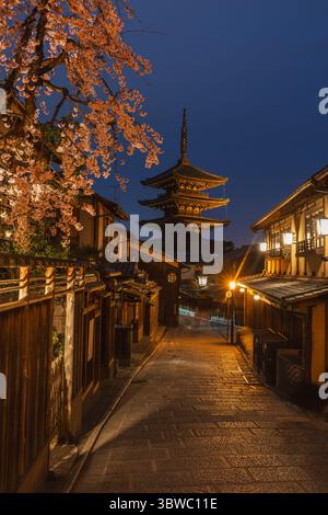 Nächtlicher Blick auf die Yasaka-Pagode in Kyoto, Japan, beleuchtet unter einem Dämmerungshimmel. Kirschblüten bilden den Rahmen für die Szene und verleihen dem Frühling Eleganz Stockfoto