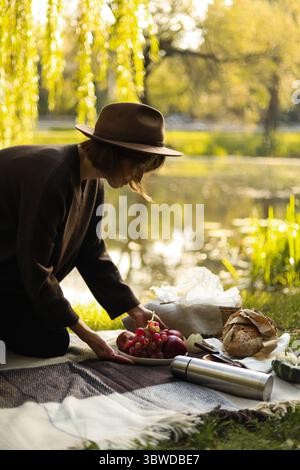 Eine junge Frau mit Filzmütze arrangiert ein Herbstpicknick in der Nähe eines friedlichen Sees und bereitet auf einer gemütlichen Decke Essen und Dekor zu. Stockfoto
