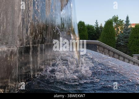 Nahansicht des Wassers, das über einen polierten Steinbrunnenrand inmitten üppiger grüner Morgenbäume fließt. Stockfoto