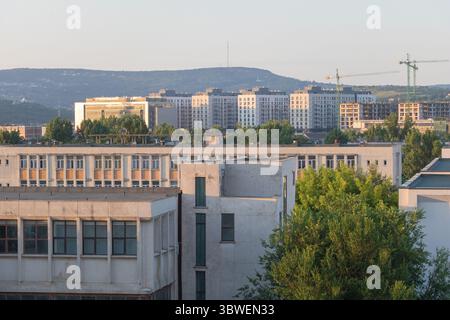 Sonnendurchflutete Skyline der Stadt mit neuen Wohnhochhäusern und Turmkränen vor hügeliger Kulisse. Stockfoto