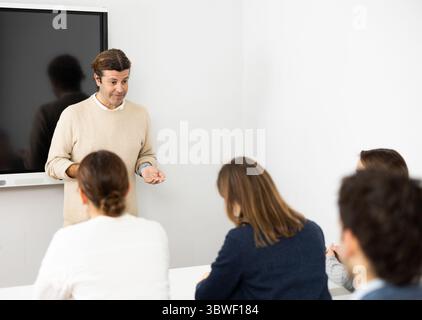 Männlicher Lehrer mittleren Alters nahe interaktivem Brett, der der Gruppe von Schülern im Klassenzimmer Regeln erklärt. Konzept des Fremdsprachenlernens Stockfoto
