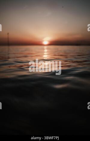 Das Bild fängt einen wunderschönen Sonnenuntergang über dem Meer ein, während die Sonne auf dem Wasser reflektiert. Der Himmel ist eine Mischung aus warmen Farben, die eine ruhige und erbsenfreie Atmosphäre schaffen Stockfoto