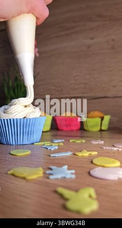 Handputzcreme auf einen Cupcake in einem blauen Etui mit farbenfrohen Hüllen im Hintergrund. Dekorative Streusel verstreut auf einem Holztisch Stockfoto