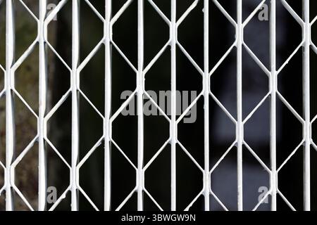 Nahaufnahme eines weißen Metalltors oder Gitters mit diamantförmigen Öffnungen vor einem dunklen, unscharfen Hintergrund. Stockfoto