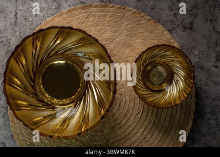 Blick von oben auf elegante goldene Schüsseln auf einem gewebten Tischset. Eine flache Komposition aus luxuriösem Geschirr auf grauem Betonhintergrund. Stockfoto