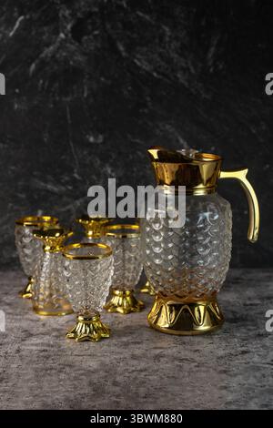 Ein Glaskrug- und Becherset im Vintage-Stil. Verziertes, strukturiertes Geschirr mit goldenen Besatz für luxuriöses Essen auf dunklem, strukturiertem Hintergrund. Stockfoto