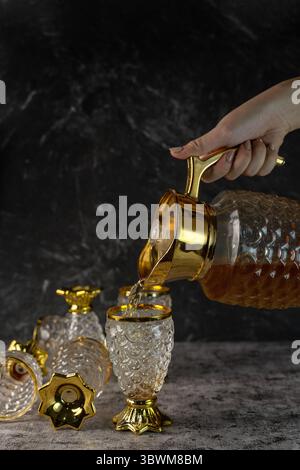 Ein Getränk aus einem alten Glaskrug in einen passenden Becher gießen. Vergoldetes Geschirr vor dunklem Marmorhintergrund. Stockfoto