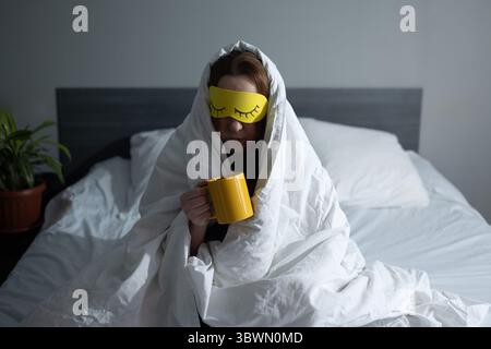 Eine wütende schlaflose Frau sitzt mit einem Becher in der Hand im Bett, sie ist wütend, weil sie früh am Morgen aufstehen muss Stockfoto