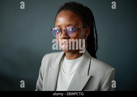 Porträt einer jungen erwachsenen Schwarzen Frau, die Brillen und Schmuck trägt und mit selbstbewusstem Ausdruck in die Kamera blickt, in professioneller Kleidung vor einem einfachen Hintergrund steht Stockfoto