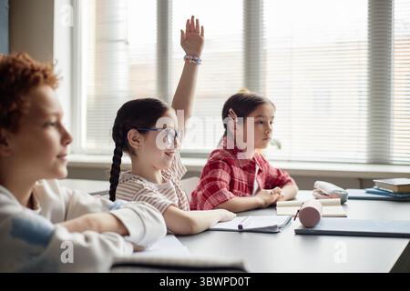 Drei Kinder sitzen am Klassenschalter, ein weißes Mädchen mit Brille, die Hand zum Lächeln hebt, zwei weitere Kinder, ein kaukasischer Junge und ein asiatisches Mädchen, die aufmerksam zuhören Stockfoto