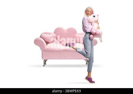 Glückliche Frau, die ein großes rosafarbenes Einhorn umarmt, vor einem rosa Sofa, isoliert auf weißem Hintergrund Stockfoto