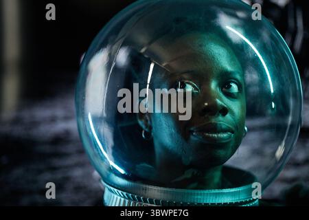 Porträt einer jungen erwachsenen Schwarzen Frau, die in einem transparenten Astronautenhelm nach oben blickt, Gesicht von blauem Licht beleuchtet, was auf futuristisches oder Weltraumforschung hindeutet Stockfoto