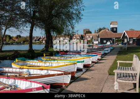 Farbenfrohe Ruderboote liegen bei Thorpeness Meare in Suffolk, England. Im Hintergrund schwimmen Schwäne auf dem Wasser. Häuser auf der gegenüberliegenden Bank. Stockfoto