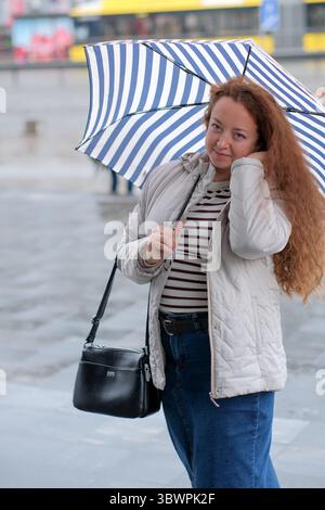 Lockige Frau genießt einen leichten Nieseln, während sie einen stilvollen Regenschirm hält. Stockfoto