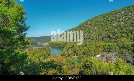 Aus der Vogelperspektive auf den Fluss Krka, der sich durch üppige grüne Hügel und bewaldete Landschaft im Krka-Nationalpark, Kroatien, unter einem hellblauen Himmel schlängelt. Stockfoto