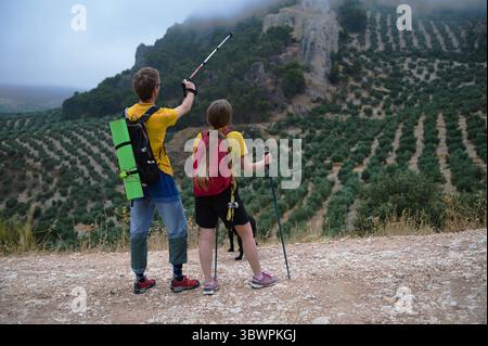 Ein junges Paar wandert durch eine malerische Berglandschaft mit Olivenhainen und trägt Trekkingausrüstung. Das Bild erinnert an Erkundungen, Naturverbundenheit, Stockfoto