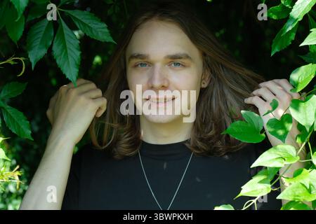 Ein junger Erwachsener steht in lebendigem Laub und strahlt Glück aus, während er sanft die Blätter berührt. Stockfoto