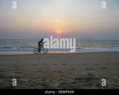 Mann, der bei Sonnenuntergang am Strand entlang fährt, mit ruhigen Meereswellen und weichem Pastellhimmel im Hintergrund. Stockfoto