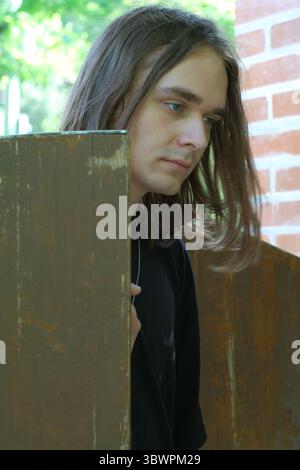 Ein junger Mensch mit langen Haaren steht nachdenklich hinter einem rustikalen Holztor. Stockfoto
