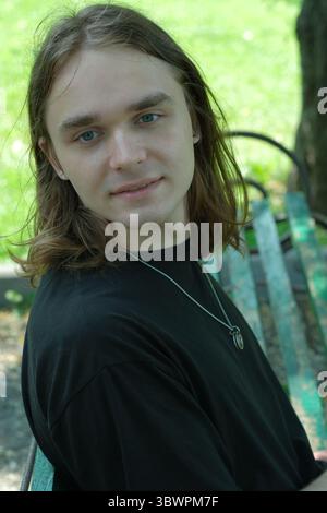 Ein junger Mann mit langen Haaren sitzt auf einer Bank in einem grünen Park, tief in Gedanken. Stockfoto