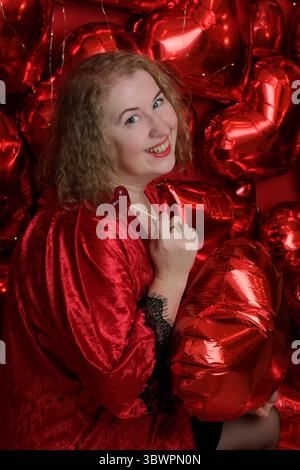Eine ältere hübsche blonde lockige Frau in einem verspielten roten Gewand posiert im Studio mit Herzballons. Stockfoto