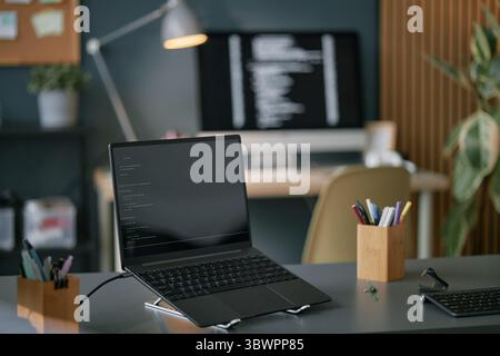 Moderner Arbeitsbereich mit offenem Laptop mit Code im Vordergrund, Desktop-Monitor mit Programmcode im Hintergrund, organisierter Schreibtisch mit sichtbaren Büromaterialien Stockfoto