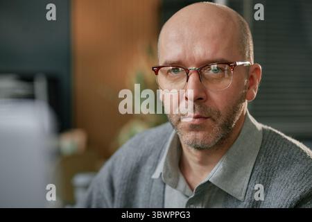 Porträt eines kaukasischen Mannes mittleren Alters, der eine Brille trägt und direkt in die Kamera blickt, mit ernsthaftem Ausdruck, der drinnen im Büro sitzt und am Computer arbeitet Stockfoto