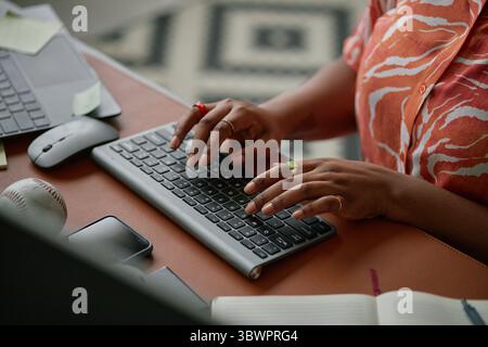 Schwarze Frau, die auf der drahtlosen Tastatur am Schreibtisch schreibt, Hände mit Ringen sichtbar, in der Nähe von Laptop, Smartphone, Notebook und Baseball arbeitet, was Multitasking in modernen Arbeitsbereichen vorschlägt Stockfoto
