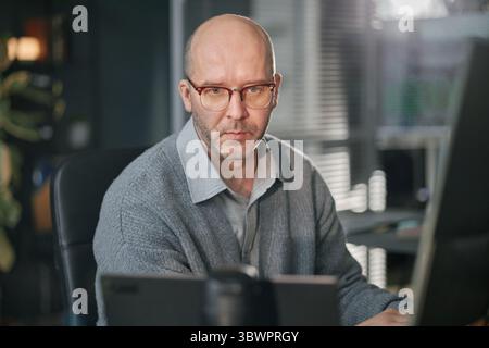 Porträt eines kaukasischen Mannes mittleren Alters mit einer Brille, der am Schreibtisch sitzt und am Computer arbeitet, in einem modernen Büro mit Blick auf den Bildschirm, kurze Haare und Stoppeln sichtbar Stockfoto
