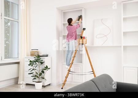 Frau, die zu Hause Bücher in Regale legt, indem sie Leiter benutzt Stockfoto