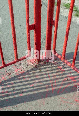 Nahaufnahme eines verwitterten roten Metallzauns, der komplizierte Schatten auf den Asphalt wirft und Anzeichen von Verschleiß im Laufe der Zeit offenbart Stockfoto