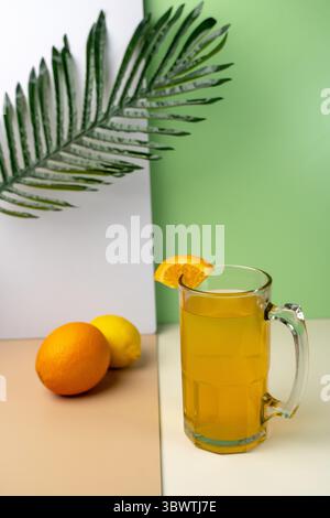 Erfrischender Orangensaft in einem Glasbecher mit einer Scheibe Garnitur. Frische Zitrusfrüchte und ein Palmblatt auf einem farbenfrohen minimalistischen Hintergrund. Sommer-Getränkekonzept Stockfoto