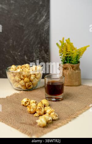 Schüssel süßes Karamell-Popcorn mit einem Glas dunkler Soda auf einer Sackleinen-Matte. Gelbe Solidago-Blüten im Hintergrund. Perfekter Snack für Filmabend. Stockfoto