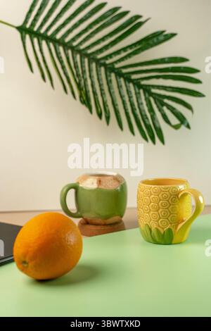 Zwei lustige, tropische Tassen in Form einer grünen Kokosnuss und einer gelben Ananas. Frische Orange und ein Palmblatt sorgen für einen Sommerurlaub. Stockfoto