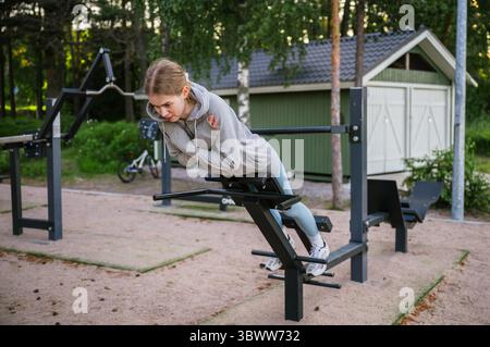 Junge Frau, die Rückenausdehnungsübungen auf der Outdoor-Fitnesbank im Sommerpark macht Stockfoto