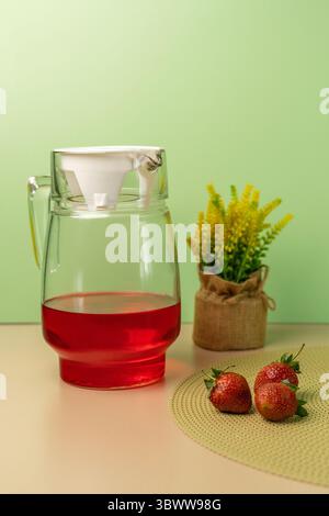 Glaskrug aus rotem Erdbeereistee oder Saft mit frischen Erdbeeren und gelben Solidago-Blüten auf grünem Hintergrund. Stockfoto