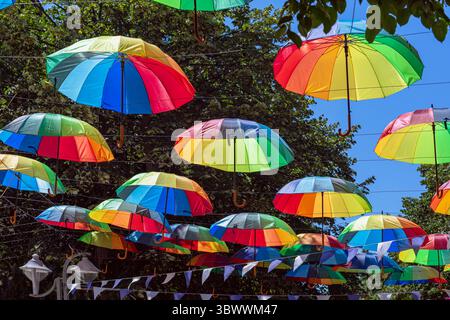 Hängende Regenbogenschirme in der Stadt. Farbenfrohe Regenschirme hängen über der Straße als künstlerische Installation, die festliche Stimmung und Schatten schafft. Konzept von Stockfoto