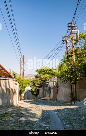 Ruhige bergauf Gasse im Bezirk Nadzaladevi in Tiflis, Georgien, mit rustikalen Häusern, Versorgungsmasten mit verwickelten Drähten und klarem blauen Kaukasus-SK Stockfoto