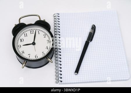 Weißes leeres Notebook mit schwarzem Wecker und Stift auf weißem Hintergrund. Bildungskonzept. Blick von oben Stockfoto