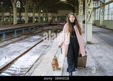 Leeres Empfangsgebäude. Junge Passagierin steht mit Koffer und kleinem Hund im Bahnhofsgebäude auf dem Bahnsteig. Wanderurlaub. Transport passend Stockfoto