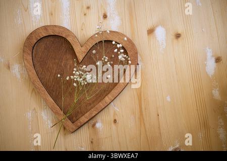 Das hölzerne Herz mit einem Zweig kleiner weißer Blumen liegt auf einem hölzernen Hintergrund Stockfoto