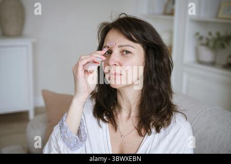 Porträt einer kaukasischen Frau, die Gesichtsmassage mit Quarzschaber für rosa Gouache macht, trägt einen Bademantel, der auf Sofa im gemütlichen Wohnzimmer zu Hause sitzt, Anti Stockfoto