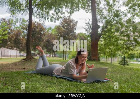 Frau, die draußen liegt grüne Yogamatte Gras lächelndes silbernes Laptop offen kommunizierende erhöhte Hand blasses Hemd graue Leggings grüner Park Hintergrund. Stockfoto