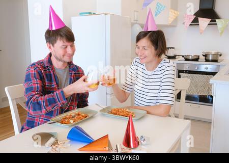 Das junge Paar feiert zu Hause gemütlich Geburtstag und genießt Kuchen und Lachen in warmer Atmosphäre. Die Familie isst Pasta und trinkt Wein, um zu feiern, zu essen und zu jubeln Stockfoto