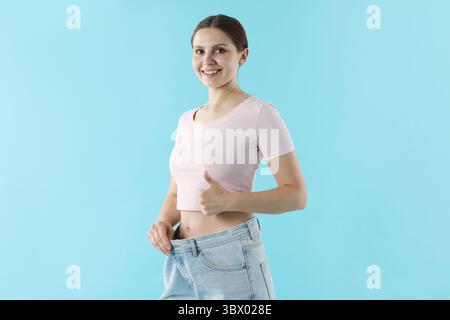Schöne Frau, die alte Jeans nach dem Abnehmen trägt und Daumen auf hellblauem Hintergrund zeigt Stockfoto
