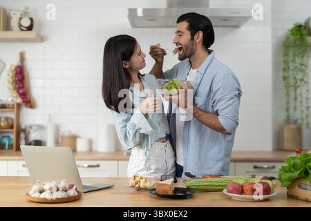 Junges Paar genießt gesundes Essen zu Hause. Ein lächelnder Mann schmeckt frischen Salat, während die Frau in der modernen Küche mit Obst die Daumen nach oben zeigt Stockfoto