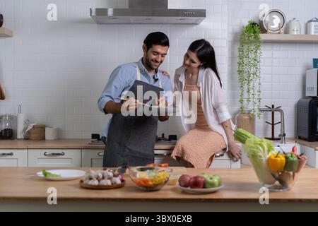 Ein junges Paar genießt einen glücklichen Kochmoment in der modernen Küche. Ein Mann in Schürze bereitet Essen zu, während die Frau vor Liebe lächelt. Frisches Obst und Gemüse Stockfoto