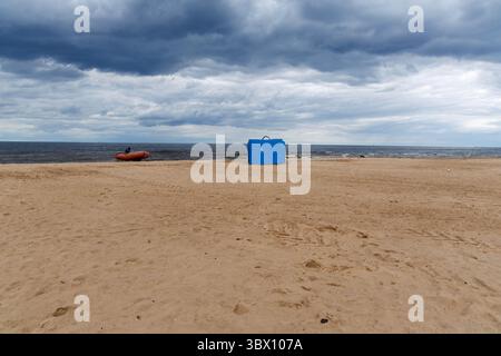 Ein blauer Rettungsschwimmer und ein aufblasbares Boot sitzen an einem breiten Sandstrand unter einem dramatischen bewölkten Himmel, in der Nähe der ruhigen Ostsee. Stockfoto
