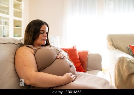 Schwangere Frau sitzt auf einem Sofa mit Händen auf dem Bauch und genießt tagsüber einen ruhigen Moment in einem natürlich beleuchteten Wohnzimmer. Lifestyle. Pregnan Stockfoto